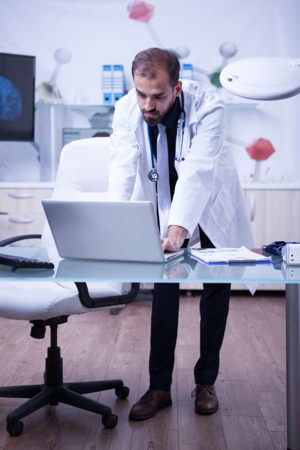 Full lenght portrait of handsome doctor working on his laptop at the hospital cabinet.